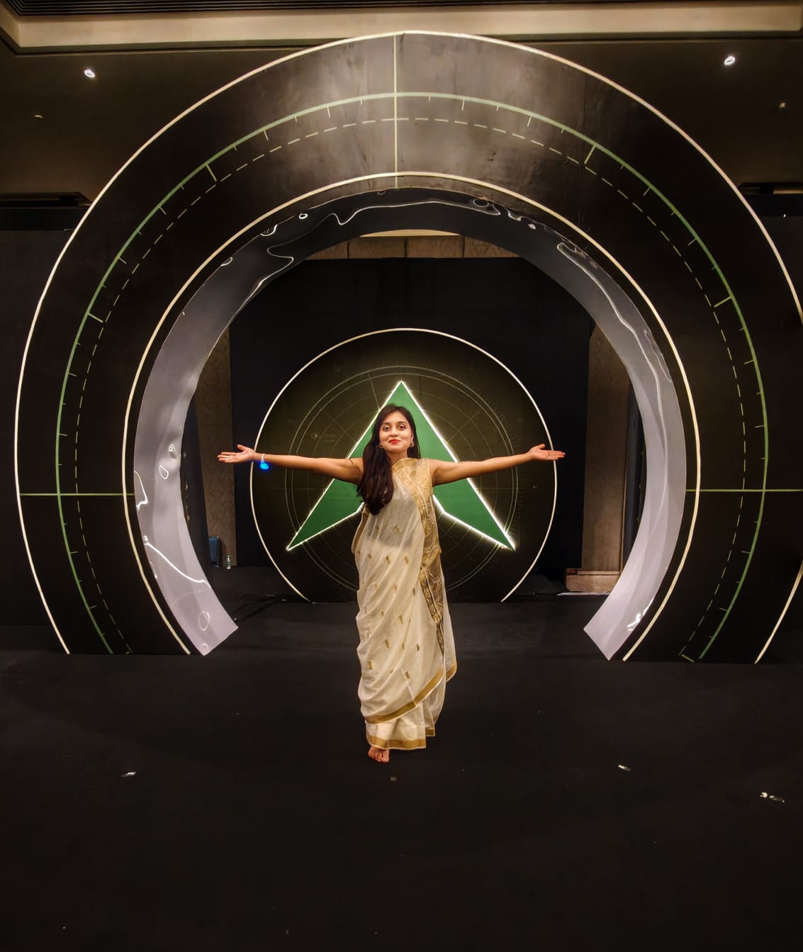 Dr Kuheli Bhattacharya standing on stage with open arms, wearing a cream saree with a gold border